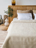 A beige sheep wool blanket is draped over a bed with white pillows in a warm, rustic bedroom setting featuring a wooden headboard, a woven pouf, and a vase.