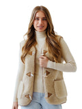 A woman modeling a beige sherpa woolen modern vest with brown leather trim and toggle closures over a cream ribbed turtleneck sweater and light blue jeans, on a white background.