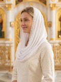 A woman wearing a white embroidered dress and a matching white headscarf with lace trim, standing in a brightly lit church interior with gold and white architectural details.