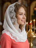 A woman wearing a white lace-trimmed veil over her head and shoulders, paired with a coral-pink top, photographed indoors in a softly blurred church-like setting with lit candles.