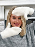 A smiling blonde woman wearing white knit woolen fluffy  mittens and a gray sweater, posing indoors with her hands raised near her face.