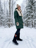 A woman standing in a snowy forest wearing a dark green winter parka with a cream sherpa collar, a beige knit beanie, knit mittens, and black rubber boots. A pair of traditional deep black Slavic valenki felt boots standing upright side-by-side in deep, fresh white snow. The boot in the foreground features a beautiful, bright red embroidery pattern on the outer side of the shaft, illustrating a traditional bunch of rowan berries hanging from a golden branch with a detailed red leaf. 