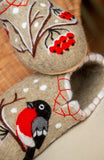 Close-up of extra comfortable felted wool slippers displaying a thick. The pristine white felt surfaces serve as a striking canvas for a vibrant red and black bullfinch bird and rowan berry arrangement hand-stitched near the foot openings.
