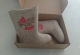 Slavic handwoven woolen boots with high shaft and without outsole in a craft box.  Overhead close-up shot of artisanal Slavic-style winter boots crafted entirely from light beige natural wool felt, resting inside an open kraft paper box. One boot stands upright, showing off a traditional folklore-inspired red berry and floral embroidery pattern on the leg section. The other boot lies flat inside the box, demonstrating its classic soleless design.
