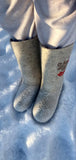Slavic high shafted woolen boots without outsole with rowanberry embroidery on a woman`s legs outdoor on the snow. The boots feature a distinct heather gray textured wool fabric with an elegant red and brown berry vine pattern embroidered on the side of the shaft. The front-facing boot is adorned with a delicate traditional embroidery design depicting red rowan or guelder-rose berries, bringing an authentic heritage look to this seamless cold-weather footwear.