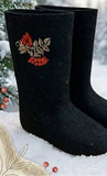 A pair of black felt booties with red and gold embroidered berry and leaf motifs, displayed in a snowy outdoor setting with pine branches. A close-up view of artisanal black felt boots highlighting the textured, dense 100 percent natural wool construction. The boots are styled without an attached outsole, showcasing a classic soft felt bottom that seamlessly merges with the rounded toe box and tall, wide leg opening. 