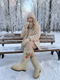 A Slavic-style woman wearing winter valenki for women with a modern protective weatherproof bottom sole, decorated with folk art embroidery of red berries and green leaves. Premium light beige felted wool boots with an integrated frost-proof outer sole, displayed in a picturesque winter landscape.