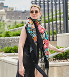 A woman wearing a very large winter stole made of natural cashmere wool over her neck for a cozy wrapping, showcasing a rich pattern of vintage peonies and blue wildflowers with yellow centers, finished with a black fringed hem.