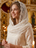 A woman wearing a white embroidered lace head covering and matching cream-colored blouse, holding a lit candle in a warm, ornate church interior.