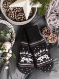 A pair of black knit winter mittens with a white Nordic-style pattern of reindeer, stars, and snowflakes, styled in a cozy flat-lay with pinecones, a star ornament, and a knitted gray scarf on a dark gray fabric background.