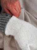 Close-up of a person's hand adjusting a soft white knit sleeve or mitten over a gray ribbed knit garment, with a beige textured blanket in the background.