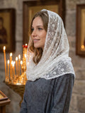A woman wearing a white lace head covering and a gray patterned dress, standing indoors in front of a stone wall with lit candles in the background.