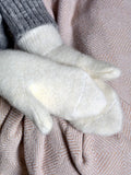 A close-up of white-colored knit mittens resting on a soft pink textured blanket, with a gray ribbed sleeve visible at the top left.