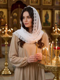 A woman in a beige dress and white lace-trimmed headscarf stands inside a candlelit church or monastery, holding a lit candle. Religious icon paintings and gold candle holders are visible in the background.
