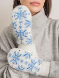 A close-up of a person wearing white knit woolen mittens with blue snowflake patterns, paired with a light gray turtleneck sweater.