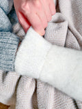 Close-up of a person's hand pulling open a white fuzzy mitten on a light beige patterned blanket, with a blue-gray knit sleeve visible on the left.