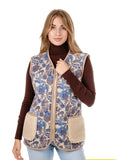 A woman modeling a sleeveless sheep wool vest with a blue and beige floral pattern and beige sherpa lining, worn over a brown turtleneck and light blue jeans against a white background.