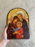 A hand holds a hand-painted wooden icon of the Holy Family (Mary, Joseph, and baby Jesus) against a textured light gray background. Hand-carved look religious wall plaque of Saint Joseph, Virgin Mary, and Baby Jesus. The detailed craquelure texture brings historical monastery reverence into a modern home altar.