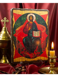 A religious icon plaque of Jesus Christ seated on a throne, displayed on a red satin drape with a brass chalice and a lit candle in the background. The icon is also well-known as Christ in Majesty.