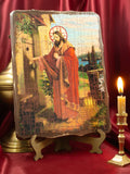 A tabletop Christian icon titled "Christ Knocks on the Door" made on a varnished pine wood board with an aged crackle finish. The artwork depicts Jesus Christ with a halo and a red cloak, standing before a scenic southern landscape as he knocks on a wooden door with no outside handle. The icon rests on a stand atop a red cloth next to a lit candle in a bronze holder.