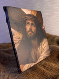 A rectangular canvas print featuring a painted portrait of Jesus carrying the cross, displayed standing upright on brown burlap fabric against a neutral textured background. Devotional Catholic artwork of Christ on the Via Dolorosa with a textured faux-gesso finish. Close-up of a rustic wood block icon featuring the face of Jesus looking up toward heaven.