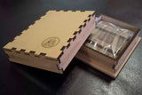 A light brown wooden presentation box with interlocking puzzle-style edges, shown closed with a circular embossed logo on the lid, next to an open box containing a clear plastic-wrapped icon, on a dark surface.