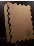 A square tan cardboard box with interlocking cut edges, photographed on a dark studio background. A circular black printed emblem appears near the lower right corner of the front face.
