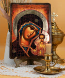 The brown wooden icon depicts a close-up portrait of the Virgin Mary (Theotokos) looking out with a somber, contemplative expression. Her head is tilted slightly toward the young Christ Child, who stands on the right. Mary wears a deep blue veil with a golden trim under a dark, textured brown or crimson maphorion (outer robe). A large, textured gold halo surrounds her head.