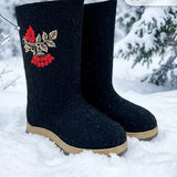 A pair of tall black felt winter boots with red berry and gold leaf embroidery, standing in snow outdoors. High-end lifestyle presentation of unique, one-of-a-kind black felted wool boots with integrated rubber outsoles for winter weather. The boots are lightly dusted with fresh white snow as they rest on the slats of a rustic wooden bench in a birch forest.