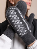A close-up of a person wearing dark gray knit woolen mittens with a white Nordic-style geometric pattern. The person is also wearing a white top and a black jacket.