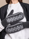 A close-up of a person wearing dark gray knit premium quality woolen mittens with a white Fair Isle snowflake pattern, layered over a white sweatshirt and black quilted jacket.