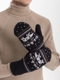 A close-up of a person wearing beige ribbed knit turtleneck sweater and holding a pair of black knit winter mittens with white snowflake and star patterns and a polar bear.