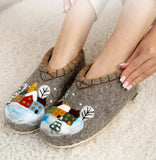 Gray warm felted slippers with winter houses and trees in snow scene design worn by a person on a light carpet.