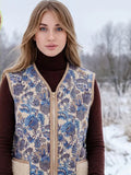 A woman wearing a sleeveless floral-patterned vest with beige sherpa lining sits on horseback outdoors in a snowy landscape. A yellow inset shows a close-up of the wool-like material.