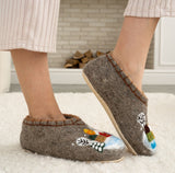 Brown merino wool slippers with decorative houses designs worn by a person indoors.